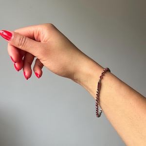 May include: A delicate bracelet with small, round, deep red beads. The bracelet is worn on a wrist with red nail polish. The background is a solid, neutral grey.