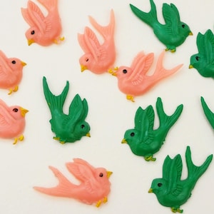 May include: A collection of plastic bird figurines in shades of pink and green. The birds have yellow beaks and feet. The figurines are arranged on a white surface, creating a whimsical and decorative display.