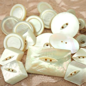 May include: A collection of various off-white buttons in different shapes and sizes. The buttons include round, oval, square, and geometric designs, with two or four holes for sewing. The buttons have a pearlescent sheen.