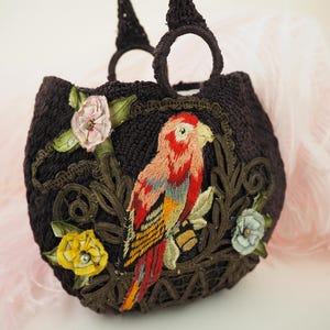 May include: A brown crocheted handbag with a parrot and floral embroidery. The parrot is red, yellow, and blue. The flowers are pink, yellow, and white.