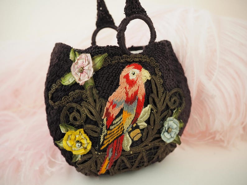 May include: A brown crocheted handbag with a parrot and floral embroidery. The parrot is red, yellow, and blue. The flowers are pink, yellow, and white.
