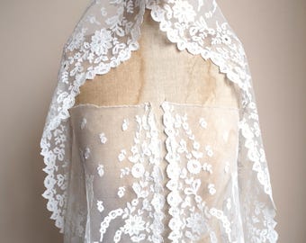 May include: A white lace veil featuring floral and leaf patterns. The veil is draped over a mannequin, highlighting its delicate design and scalloped edges. The lace is detailed with various floral motifs.