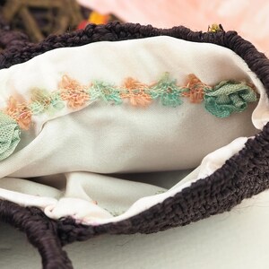 May include: A brown crocheted bag with a white fabric lining. The lining has a decorative trim of pink and green fabric rosettes.