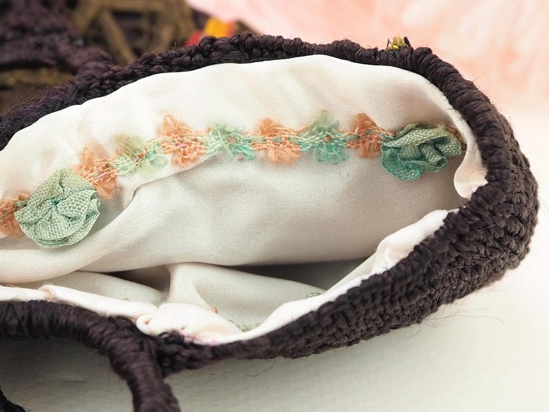 May include: A brown crocheted bag with a white fabric lining. The lining has a decorative trim of pink and green fabric rosettes.