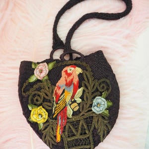May include: A small, brown, crocheted purse with a parrot embroidered on the front. The parrot is red, yellow, and blue, and is sitting in a cage with green vines and flowers. The purse has a brown crocheted strap.