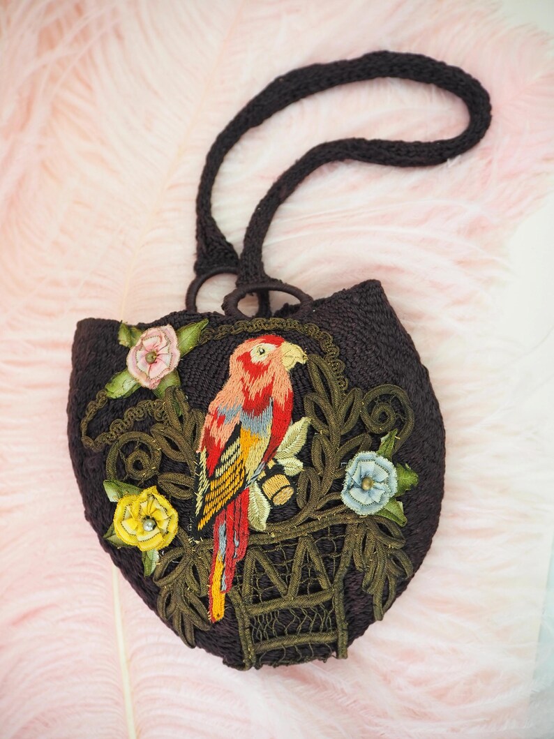 May include: A small, brown, crocheted purse with a parrot embroidered on the front. The parrot is red, yellow, and blue, and is sitting in a cage with green vines and flowers. The purse has a brown crocheted strap.