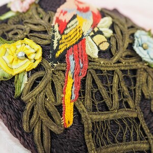 May include: A close-up of a dark brown fabric heart-shaped pincushion with gold and green embroidery. The embroidery features a bird, flowers, and a cage.