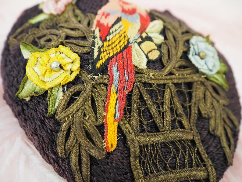 May include: A close-up of a dark brown fabric heart-shaped pincushion with gold and green embroidery. The embroidery features a bird, flowers, and a cage.