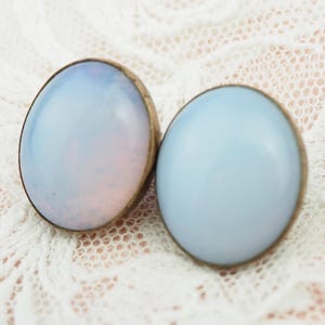 May include: A pair of vintage stud earrings. Each earring features a light blue, oval-shaped stone with a gold-tone metal setting. The earrings are displayed on a white lace surface.
