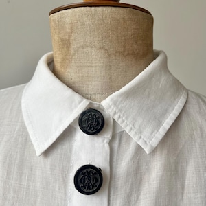 May include: Close-up of a white linen shirt with a classic collar and two large, round black buttons. The buttons feature an ornate, embossed design. The shirt is displayed on a mannequin.