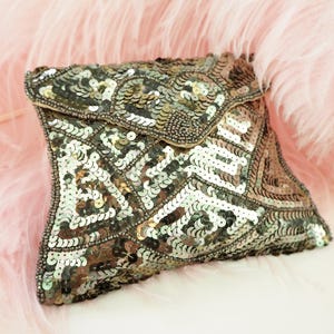 May include: A silver sequined clutch with a flap closure. The bag is covered in small, round sequins in shades of silver and gray, arranged in a geometric pattern. The background is a soft pink feather boa.