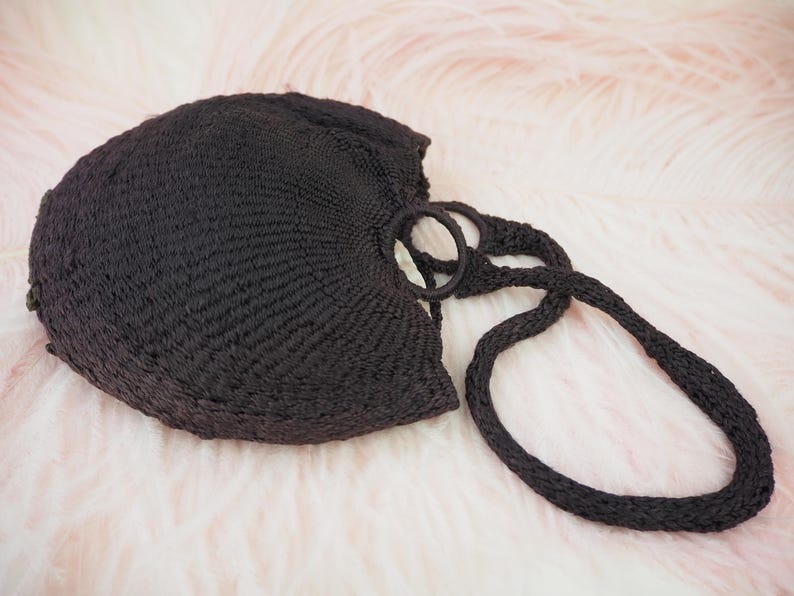 May include: A black crocheted tote bag with a single, thick strap. The bag is made of a woven material and has a round, open top.