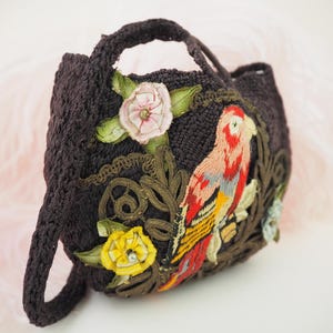 May include: A small black crocheted bag with a parrot and floral embroidery. The parrot is red, yellow, and blue. The flowers are pink and yellow.