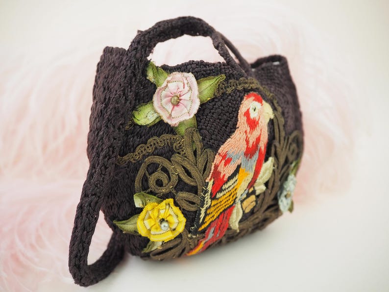 May include: A small black crocheted bag with a parrot and floral embroidery. The parrot is red, yellow, and blue. The flowers are pink and yellow.