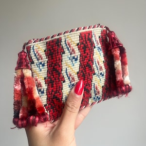 May include: A rectangular, woven clutch with a red, white, and black striped pattern. The clutch has red tassels on each end and a braided border. The clutch is held in a hand with red nail polish.