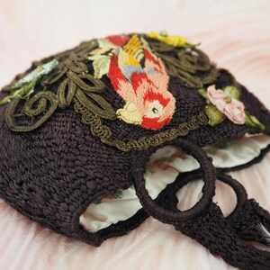 May include: A dark brown crocheted tote bag with embroidered floral and bird designs. The bag has two woven handles.