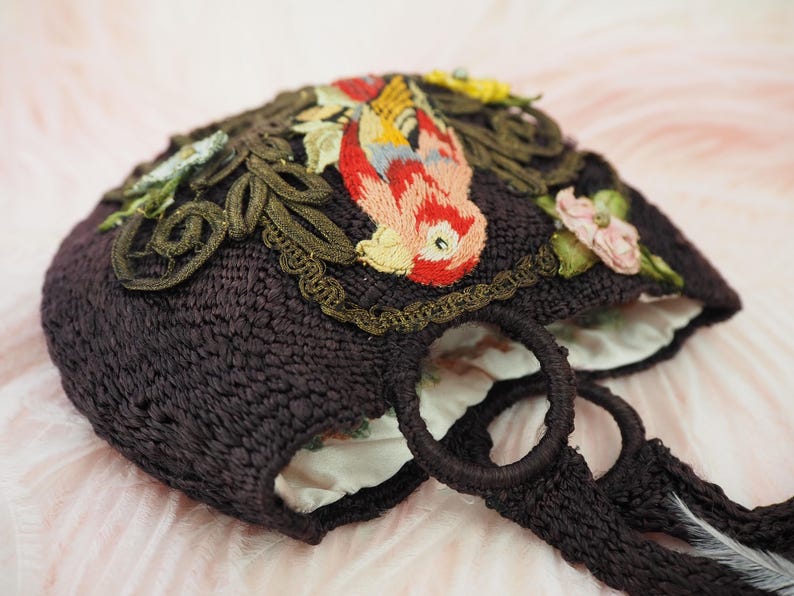 May include: A dark brown crocheted tote bag with embroidered floral and bird designs. The bag has two woven handles.