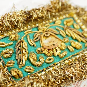 May include: A teal fabric rectangle with gold embroidery and sequins. The embroidery depicts floral and abstract patterns. The edges are trimmed with gold metallic fringe. It is likely a decorative element for clothing or home decor.