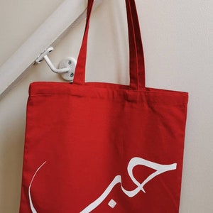 May include: A red canvas tote bag with white Arabic calligraphy that says "حب" which means "love" in English.