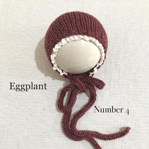 May include: A knitted baby bonnet in eggplant purple with a white lace trim and matching ties. The bonnet is labelled "Number 4".