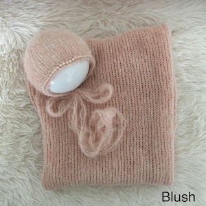 May include: A blush pink knitted baby hat and wrap set with a pearl trim on the hat. The set is laid out on a white fluffy surface.