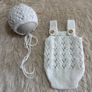 May include: A white knitted baby romper and matching bonnet set. The romper features a delicate openwork pattern, two shoulder straps with small wooden buttons, and a rounded bottom. The bonnet has a matching lace-like knit design.