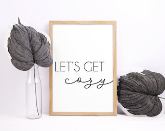 May include: A framed print with the words "LET'S GET cozy" in black lettering. The print is in a light wood frame and is next to a clear glass bottle with a black leaf and a black and white leaf.
