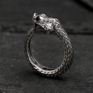 May include: A silver ring featuring a detailed snake design. The snake's head is raised and has a black eye. The ring is made of textured metal.