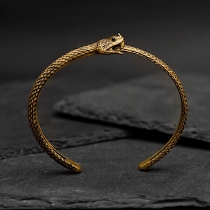 May include: A gold-toned snake bangle bracelet with a detailed snake head design. The bracelet is open and has a simple, minimalist style.