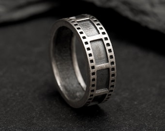 May include: A silver-toned ring featuring a band designed to resemble a strip of 35mm camera film. The exterior surface displays a repeating pattern of rectangular frames bordered by small square sprocket holes, finished with a dark, matte texture.