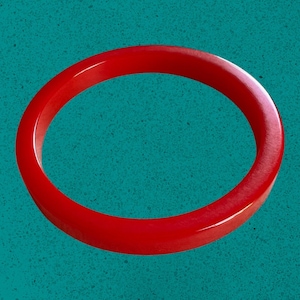May include: A vibrant red bangle bracelet. The circular bracelet is a solid, glossy red color. The background is a textured teal color.