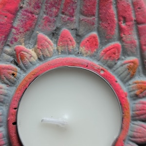 May include: A circular tea light candle holder made of gray concrete with a hand-painted pink and orange lotus flower design. The holder features a central circular cutout containing a white wax tea light candle with a visible wick.