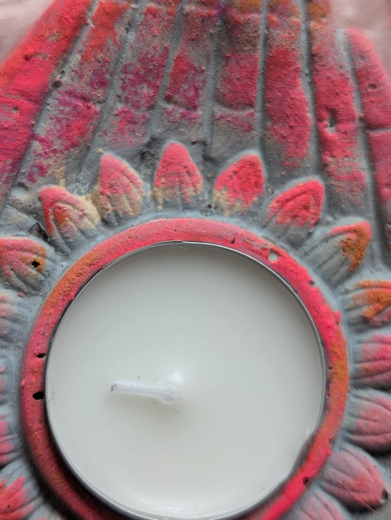 May include: A circular tea light candle holder made of gray concrete with a hand-painted pink and orange lotus flower design. The holder features a central circular cutout containing a white wax tea light candle with a visible wick.
