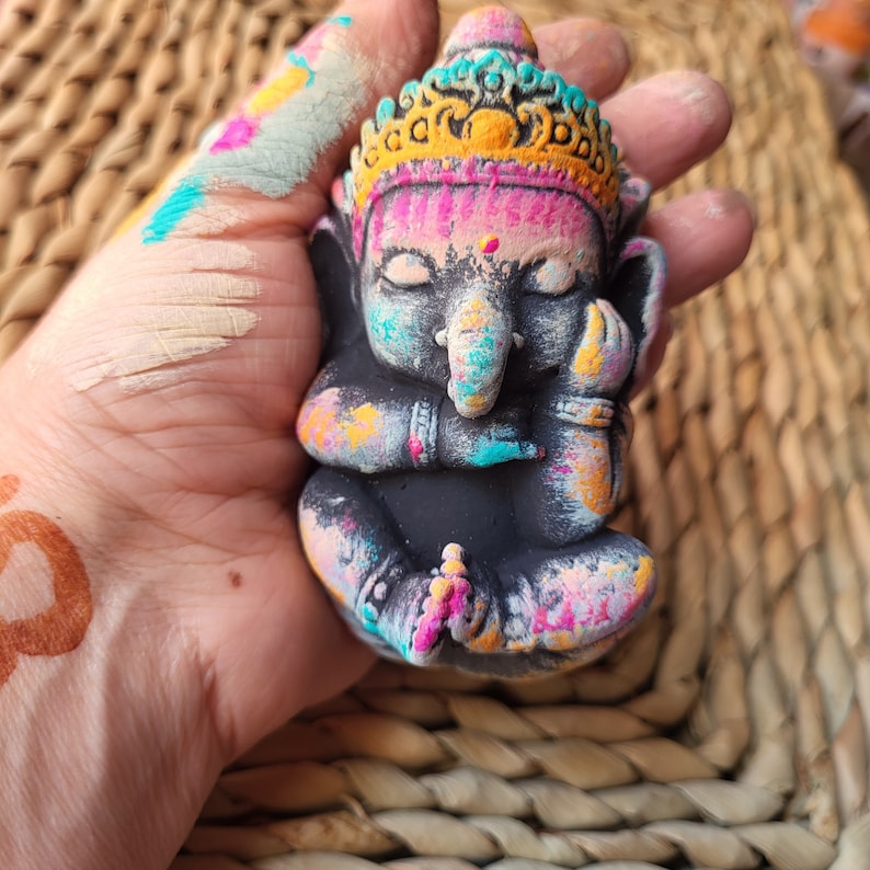 May include: A small, colourful statue of Ganesha, the Hindu god of wisdom and prosperity, with a black base and painted in shades of pink, blue, yellow, and white.