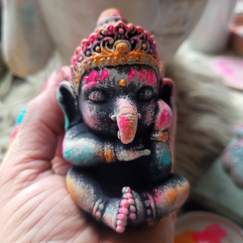 May include: A small, black painted figurine of Ganesha, the Hindu god of beginnings, with a pink and orange crown and colourful paint splatters.