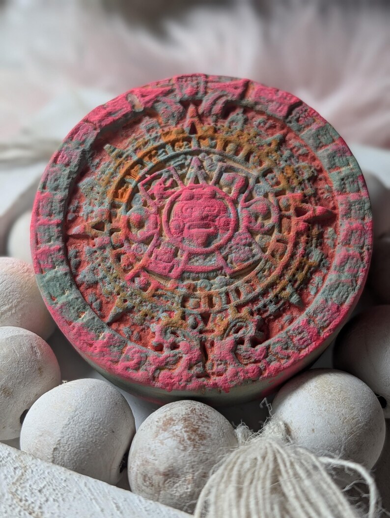 May include: A circular, flat object featuring a detailed, embossed Aztec calendar design in shades of vibrant pink, orange, and muted gray. The surface has a textured, matte appearance with intricate carvings of symbols and a central face motif.