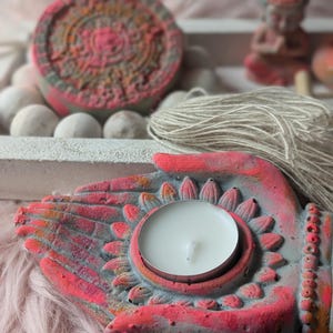 May include: A concrete candle holder shaped like two cupped hands in a grey and vibrant pink finish, holding a white tea light candle. The design features a lotus petal motif surrounding the candle base, with a textured surface and a tassel.
