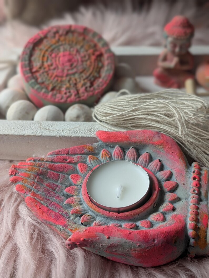 May include: A concrete candle holder shaped like two cupped hands in a grey and vibrant pink finish, holding a white tea light candle. The design features a lotus petal motif surrounding the candle base, with a textured surface and a tassel.