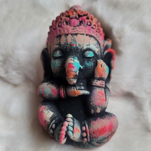 May include: A small, colourful, hand-painted statue of Ganesha, the Hindu god of wisdom and prosperity. The statue is made of a dark material and features a pink crown, blue eyes, and a colourful trunk.