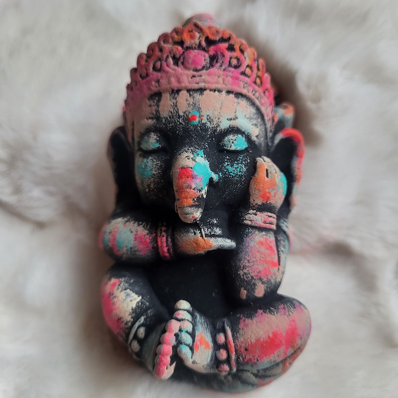 May include: A small, colourful, hand-painted statue of Ganesha, the Hindu god of wisdom and prosperity. The statue is made of a dark material and features a pink crown, blue eyes, and a colourful trunk.
