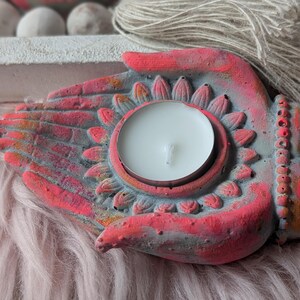 May include: A hand-shaped candle holder made of concrete, featuring a grey base with vibrant pink and orange painted accents. The center of the palm holds a circular white tea light candle, surrounded by a carved lotus petal design.
