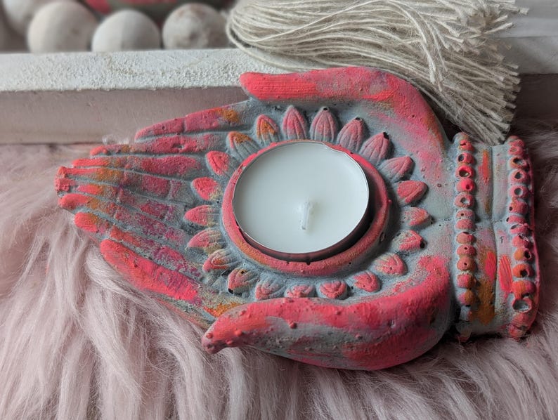 May include: A hand-shaped candle holder made of concrete, featuring a grey base with vibrant pink and orange painted accents. The center of the palm holds a circular white tea light candle, surrounded by a carved lotus petal design.