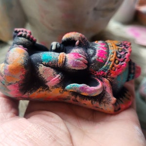 May include: A small, black, hand-painted elephant figurine with colourful accents. The elephant is in a sleeping position with its trunk curled up. The figurine is decorated with pink, orange, blue, and green paint.