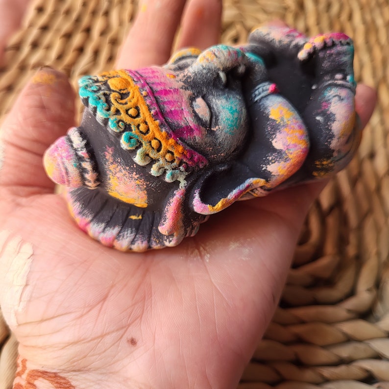 May include: A small, black, painted ceramic figurine of the Hindu deity Ganesha. The figurine is decorated with colourful paint, including pink, yellow, blue, and green. The figurine is in a sleeping position with its trunk curled up.