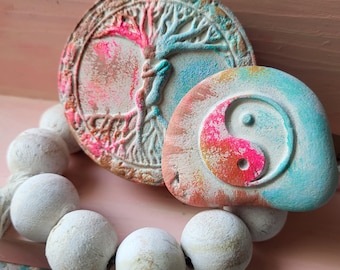 May include: A handmade bracelet featuring large, white, round beads. The bracelet is adorned with two decorative, textured, stone-like pendants. One pendant features a tree of life design, and the other a yin-yang symbol, both with colorful accents.