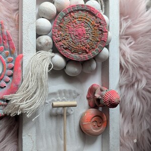 May include: A rectangular white wooden frame containing a sand garden with a wooden rake, a small red and orange Buddha figurine, a circular yin-yang stone, and a circular calendar stone surrounded by white wooden beads and a hanging tassel.