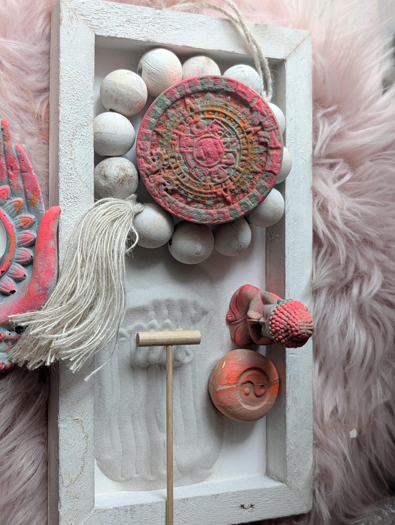 May include: A rectangular white wooden frame containing a sand garden with a wooden rake, a small red and orange Buddha figurine, a circular yin-yang stone, and a circular calendar stone surrounded by white wooden beads and a hanging tassel.