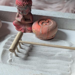 May include: A desktop zen garden set featuring a small red Buddha figurine in a prayer pose, a round orange and white marbled stone with an embossed yin-yang symbol, and a light-colored wooden rake resting on a bed of fine white sand.