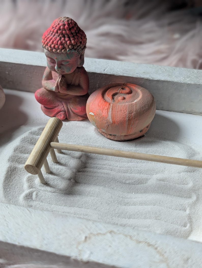 May include: A desktop zen garden set featuring a small red Buddha figurine in a prayer pose, a round orange and white marbled stone with an embossed yin-yang symbol, and a light-colored wooden rake resting on a bed of fine white sand.