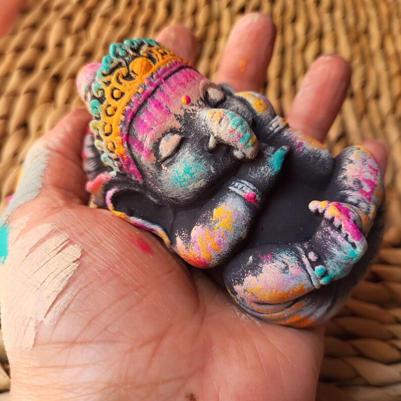 May include: A small, black, sleeping Ganesha statue with a colourful, yellow, pink, and blue paint design. The statue is holding its trunk with its right hand and has a crown on its head.